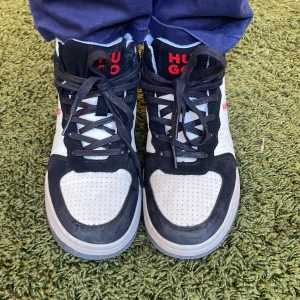 Svartvita sneakers från Hugo - Snygga svartvita sneakers från Hugo med röd logga på sidan och hälen. Skorna har snörning och en perforerad ovandel för extra ventilation. Perfekta för en stilren look.