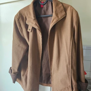 Brun jacka med perforerat mönster - Snygg brun jacka med perforerat mönster och krage. Jackan har långa ärmar och en stilren design. Perfekt för sommaren! 