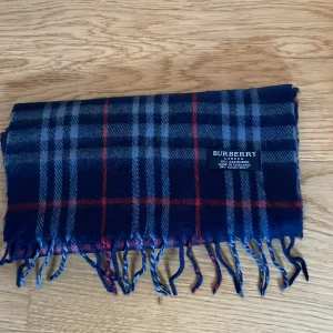 Burberry halsduk - Säljer en elegant rutig halsduk från Burberry i blått och rött. Halsduken är tillverkad i 100% kashmir och har fransar i ändarna. Perfekt för att hålla dig varm och stilren under kyliga dagar. Priset är inte hugget i sten och jag är öppen för potentiella byten. Nypris runt 4500kr