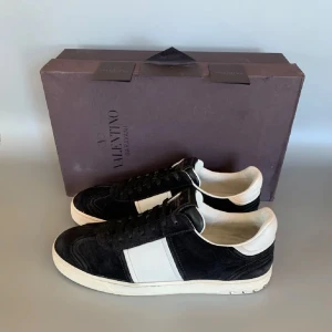 Valentino Garavani sneakers - Snygga Valentino Garavani sneakers i svart mocka med vita detaljer. Skorna har en rund tå och snörning framtill. Sulan har ett unikt mönster i grönt och svart. Bak på hälen finns de ikoniska nitarna som ger en extra edge. Helt nya skor självklart äkta! Box och kort ingår! Hör av er vid funderingar!