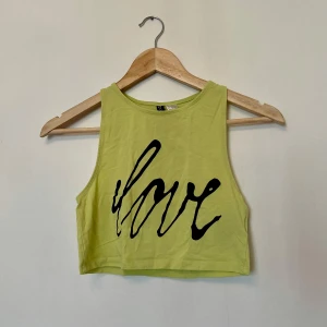 Grön croppad topp från H&M - Säljer en grön ärmlös croppad topp från H&M med texten svart på framsidan. Perfekt för varma sommardagar eller som ett lager under en jacka.
