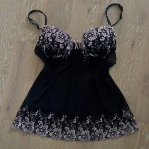 Svart blommig linnetopp - Supersöt svart linnetopp med ljusrosa broderade blommor på kuporna och längs nederkanten. Justerbara axelband och små rosetter framtill. Storleken i kuporna är 75b🥰