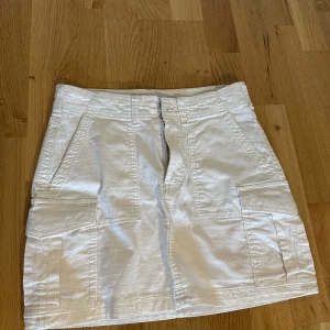 Vit mini cargo kjol från Bershka - Säljer en vit mini cargo kjol från Bershka i storlek XS. Kjolen har flera fickor både fram och bak, samt knapp och dragkedja framtill