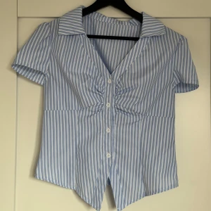 Blåvit randig skjorta med v-ringning - Kortärmad skjorta med blå och vita ränder, djup v-ringning och snygg rynkdetalj framtill. Skjortan har knappar hela vägen ner och en spetsig krage. Materialet känns som bomull och passformen är normal. Perfekt för en fräsch och stilren look.