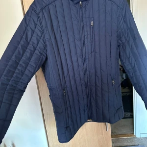 Mörkblå quiltad jacka från Signal XL - Quiltad mörkblå jacka från Signal i storlek XL. Jackan har dragkedja framtill, tre fickor med dragkedja och snyggt brunt innerfoder. Perfekt för höst och vinter, med stilrena vertikala sömmar och klassisk passform.