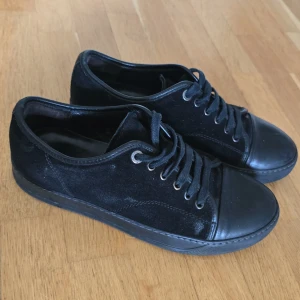 Svarta sneakers från Lanvin i mocka - Snygga svarta sneakers från Lanvin med ovandel i mocka och tå samt sula i skinn. Klassisk rund tå, svarta skosnören och diskret logga under sulan. Perfekta för dig som gillar stilrena och lyxiga detaljer i vardagen. Storlek, står 5 på tungan men sulan mäter 26,6 och motsvarar då storlek 41/42. Finns lättare skav på mockan utsida vä fot bak samt ett snöre där metalldelen i toppen lossnat. För övrigt i bra skick enligt bilderna.