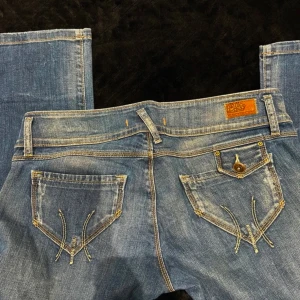 Blå jeans med fräcka fickor - Snygga blå jeans med bootcut passform och slitna detaljer. Fickorna bak har coola broderier och kontrastsömmar. Jeansen har låg midja och klassisk knappstängning. Perfekta för dig som gillar en avslappnad men trendig look.