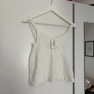 Vit spetsig linnetopp med knyt - Supersöt vit linnetopp med spetsdetaljer och knyt framtill. Toppen har breda axelband i spets och en lätt rynkad struktur som ger en romantisk vibe. Perfekt för varma dagar och somriga outfits.