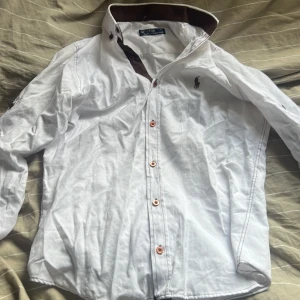 Vit skjorta från ralph lauren - Vit långärmad skjorta från ralph lauren. med klassisk krage och kontrastdetaljer i brunt på insidan av kragen. Skjortan har knappar framtill och broderad logga på bröstet. Perfekt för en clean och stilren look.