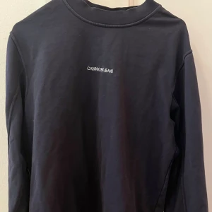 Svart sweatshirt från Calvin Klein Jeans - Säljer en svart långärmad sweatshirt från Calvin Klein Jeans med vit logotyp broderad på bröstet. Tröjan har en klassisk rund hals och är gjord i mjuk bomull, perfekt för chill dagar. Enkel och stilren design som funkar till det mesta.