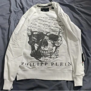 Vit sweatshirt med döskalle från Philipp Plein - Säljer en vit sweatshirt från Philipp Plein med stor döskalle framtill, dekorerad med text och glittriga detaljer. Tröjan har ribbade muddar, lång ärm och ett metallmärke med PP-logga på ryggen. Perfekt för dig som gillar edgy streetstyle.