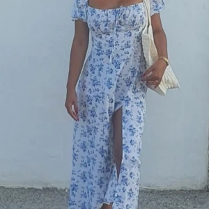 Blommig långklänning med slits - Säljer en superfin långklänning med blått blommigt mönster på vit botten. Klänningen har korta puffärmar, snörning vid bysten och en snygg slits framtill. Perfekt för varma dagar och ger en romantisk vibe.