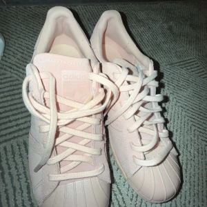 Adidas Superstar i ljusrosa mocka - Snygga Adidas Superstar sneakers i ljusrosa mocka med klassisk snäcktå och matchande ljusrosa snörning. Perfekta för dig som vill ha en stilren och trendig look med en feminin touch.