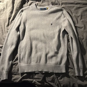 Grå stickad tröja Polo Ralph Lauren - Snygg grå stickad tröja från Polo Ralph Lauren med klassisk rund halsringning och broderad logga på bröstet. Tröjan har ribbade muddar vid ärmslut och nederkant, perfekt för en clean och stilren look.