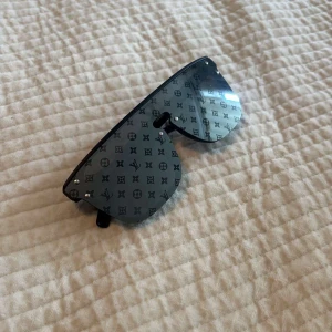 Svarta Louis Vuitton solglasögon - Säljer ett par svarta Louis Vuitton solglasögon med bred, futuristisk sköldform och monogrammönster över hela linsen. Modellen är LV2330 och har en cool, modern vibe med diskret logga på sidan. Perfekt för dig som vill sticka ut med lyxig streetstyle.