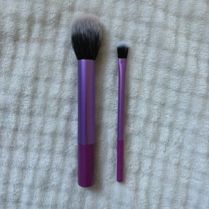 Lila sminkborstar duo - Två sminkborstar med lila och mörklila handtag samt mjuka syntetiska borst i svart och vitt. En större borste för ansiktet och en mindre för ögonskugga eller detaljer. Perfekta för att skapa en flawless bas och snygga ögonlooks.