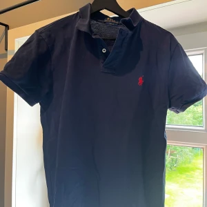 Mörkblå Polo Ralph Lauren piké - Snygg mörkblå piké från Polo Ralph Lauren i slim fit-modell. Klassisk pikékrage med två knappar och röd broderad logga på bröstet. Tillverkad i mjuk bomull som känns skön mot huden. Perfekt för dig som gillar stilrena och tidlösa plagg.