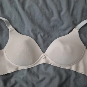 Beige axelbandslös bh med spetskant - Säljer en beige axelbandslös bh med lätt vadderade kupor och dekorativ spetskant längs nederdelen. Bh:n har avtagbara axelband och en liten silverfärgad detalj framtill. Perfekt under toppar och klänningar med bara axlar.