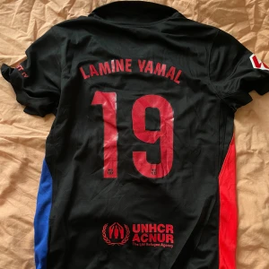 Barcelona Lamine Yamal tröja - Snygg FC Barcelona fotbollströja med Lamine Yamal och nummer 19 på ryggen. Svart bas med röda och blå sidopaneler, röda tryck och detaljer. Nike Dri-FIT material för skön känsla och bra ventilation. Spotify-logga på bröstet och UNHCR-tryck nedtill.