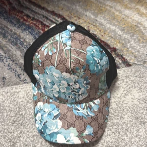 Keps - Keps med  stora blå blommor. Kepsen har svarta meshpaneler på sidorna och justerbar rem bak med klassiska färger. Perfekt statement-piece för dig som vill sticka ut. PRIS KAN DISKUTERAS  