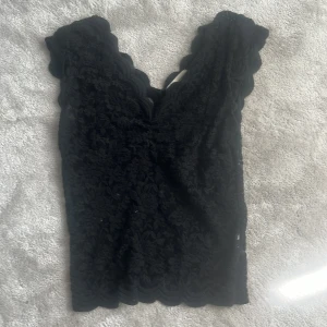 Svart spetsblus med v-ringning - Supersnygg svart blus i spets med v-ringad hals och vågiga kanter. Ärmlös modell som sitter tajt och har blommigt spetsmönster över hela toppen. Perfekt att styla med kjol eller jeans för en trendig look.