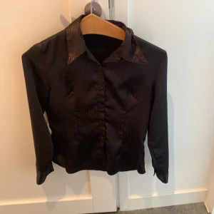 Svart randig skjorta i satin-look - Snygg svart skjorta med subtila vertikala ränder och glansig satin-känsla. Skjortan har klassisk krage, knäppning framtill och långa ärmar. Perfekt för dig som vill ha en stilren men ändå lite extra look.
