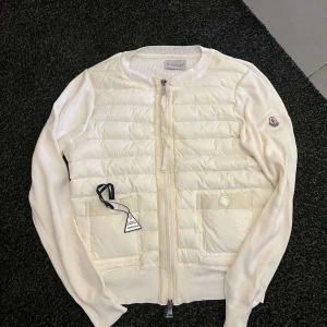 Vit jacka från Moncler - Snygg vit jacka från Moncler med quiltad framsida och två stora fickor. Västen har rund halsringning, dragkedja framtill och Moncler-logga på ärmen. Materialmix med dun och stickade partier ger en modern look.