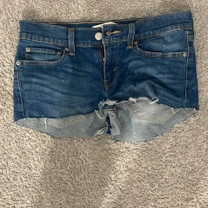 Levi's jeansshorts med rå kant - Snygga blå jeansshorts från Levi's med klassisk femficksdesign och rå, fransig kant nedtill. Shortsen har normal passform, knapp och dragkedja framtill samt tydliga Levi's-detaljer på bakfickorna och läderpatch i midjan. Perfekta för sommaren! De är ett par jeans från början som jag har klippt 
