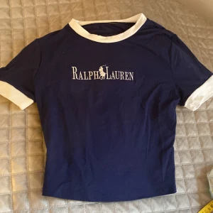 Blå Ralph Lauren t-shirt med vit kant - Snygg marinblå t-shirt från Ralph Lauren med vit broderad logga på bröstet. T-shirten har vit kant vid halsringning och ärmslut, samt klassisk rund hals. Perfekt för dig som gillar stilrena märkesplagg och enkel design.
