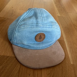 Ljusblå jeanskeps från H&M - Snygg ljusblå keps från H&M i jeansmaterial med beige mockaliknande skärm. Framtill finns en rund brun patch med texten 'Wood Creek Park' och en gran. Kepsen har elastisk bakdel för skön passform. Perfekt för att ge din stil en chill vibe.