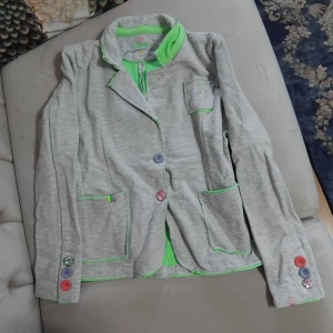 Grå kavaj med färgglada knappar Raw DNM - Unik grå kavaj från Raw DNM med gröna detaljer och broderier. Flera färgglada knappar i lila, rosa och rött på ärmar och framtill. Kavajen har tre fickor och coola gröna sömmar som sticker ut. Materialet är mjuk bomullsmix har aldrig använts men har tyvärr inte kvittot till den