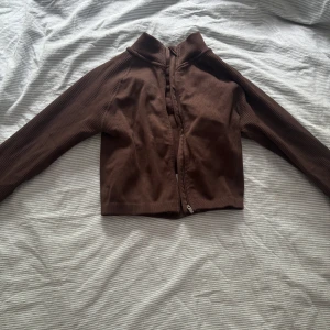 Brun ribbad zipjacka från Bo+Tee - Snygg brun ribbad jacka från Bo+Tee med croppad passform och dragkedja framtill. Jackan har långärmade ärmar och en sportig look, perfekt att slänga på sig över träningskläder eller till jeans. Materialet är stretchigt och mjukt för extra komfort.