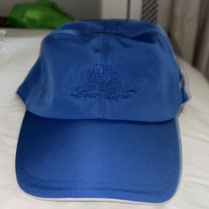 Loro piana keps - Snygg blå keps från Loro Piana Storm System med broderad logga framtill och metallmärke på sidan. Insidan har ett rutigt foder i beige, brunt och grönt. Kepsen har klassisk form och är tillverkad i ett slitstarkt syntetmaterial. Helt oanvänd då jag inte gillade passformen