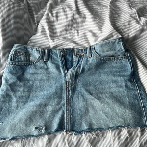 Blå jeanskjol med rå kant - Säljer en ljusblå jeanskjol med fransig, rå kant och slitna detaljer. Kjolen har klassiska fickor fram och bak, bälteshällor och knappstängning. Perfekt för dig som gillar en chill och avslappnad stil. Storlek 164 
