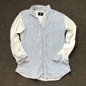 D&G skjorta i bomull - Snygg skjorta från D&G med blåvita ränder framtill och helvita ärmar och rygg. Klassisk krage, bröstfickor med knappar och långa ärmar. Tillverkad i tunn och skön bomull, perfekt för en stilren look. 