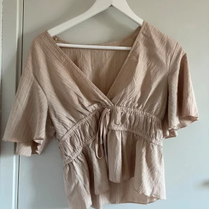 Beige omlottblus från SHEIN - Säljer en beige omlottblus från SHEIN i storlek M. Blusen har korta, vida ärmar, knytning i midjan och volangdetaljer nedtill. Materialet är lätt och har en skrynklig struktur som ger en avslappnad vibe. Perfekt för dig som gillar en feminin och trendig look. Storlek M men passar även S och Xs