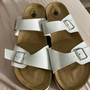 Vita sandaler med plattform och spännen - Snygga vita sandaler med breda remmar och silvriga spännen. De har en rejäl plattformsula i vitt och korkdetaljer på sidan. Ovandelen är i syntet och sulan har ett vågigt mönster undertill. Perfekta för en trendig sommarlook.