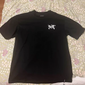 Svart Arc'teryx T-shirt - Svart t-shirt från Arc'teryx i bomull med vit logga och skelett-tryck på bröstet samt stort grafiskt tryck på ryggen. Klassisk passform och rund hals. Perfekt för dig som gillar streetwear och snygga detaljer. Helt oanvänd, endast prövad, säljer då den inte passar mig i storleken