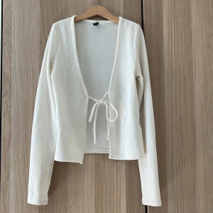 Vit ribbad kofta med knyt från H&M - Säljer en vit ribbad kofta från H&M Divided i storlek XS. Koftan har lång ärm, djup v-ringning och knytbar. Materialet är mjukt och stretchigt, vilket gör den skön att bära.