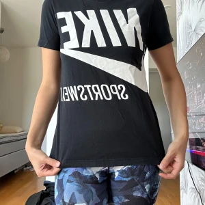 Svart Nike t-shirt med stor logga - Svart t-shirt från Nike med stor vit logga och texten 'SPORTSWEAR' på framsidan. Klassisk rund hals och korta ärmar. T-shirten är i mjuk bomull och har en normal passform som sitter snyggt och bekvämt. Perfekt för dig som gillar sportig streetstyle.