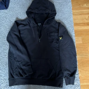 Svart hoodie från Lyle & Scott - Svart hoodie från Lyle & Scott med halv dragkedja, huva och stor ficka på bröstet. Klassisk gul logga på ärmen. Mjuk bomullskänsla och ribbade muddar. Perfekt för chill dagar och enkel att matcha med jeans eller joggers. Finns ett litet märke på ena armen som man ser i bild 4.