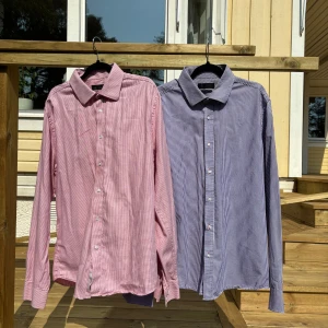 2 st Rutiga skjortor Riley Tailored Fit - Två snygga långärmade skjortor från Riley i modellen Tailored Fit. En är rödvit och den andra blåvit, båda med klassiskt rutigt mönster och knappar framtill. Skjortorna är gjorda i 100% bomull och har en stilren krage, perfekta för en clean och fräsch look.