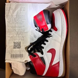 Nike Air Jordan 1 Chicago High - Nike Air Jordan 1 High i klassiska Chicago-färger. Skorna har vit bas i skinn med röda och svarta detaljer, svart snörning och röd yttersula. Ikonisk Air Jordan-logga på ankeln och extra röda skosnören medföljer. Perfekt för dig som vill sticka ut med en tidlös sneaker.