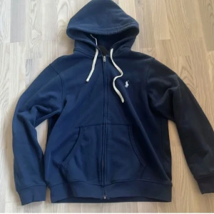 Mörkblå hoodie från Polo Ralph Lauren - Snygg mörkblå hoodie från Polo Ralph Lauren med vit broderad logga på bröstet. Tillverkad i mjuk bomull, perfekt för chill dagar eller när du vill ha en clean look.
