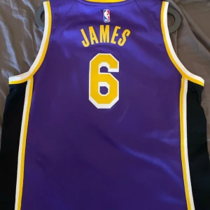 Lakers LeBron James baskettröja XL - Snygg Lakers baskettröja med LeBron James namn och nummer 6. Tröjan är lila med gula och vita detaljer, har ärmlös design och är tillverkad i Dri-Fit material som andas. Lakers-logga och NBA-märke på framsidan, perfekt för dig som älskar basket och streetwear. Storleken passar alla M-XL