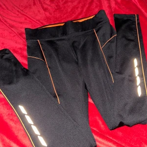 Svarta träningsleggings med reflex - Svarta träningsleggings med orangea detaljer och reflexränder på benen. Byxorna har hög midja och en liten dragkedjeficka bak. Perfekta för löpning eller gym, med snygg passform och sportig look.