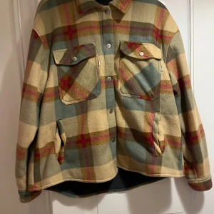 Rutig overshirt från CRW i fleece - Riktigt snygg overshirt från CRW i mjuk fleece. Jackan har rutigt mönster i beige, grönt, brunt och rosa, med knappar och två stora bröstfickor. Perfekt för lager-på-lager och chillare stil.