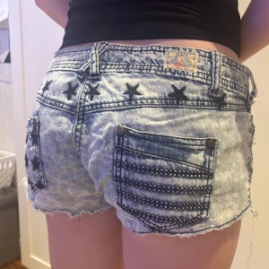Low rise jeansshorts - Midjemått: 40cm längd på kanten: 19cm Säljer ett par coola ljusblå jeansshorts från SC&CO med slitna kanter, svarta stjärnor och ränder som ger en unik look. Shortsen har klassiska fickor och låg midja, perfekt för sommaren.