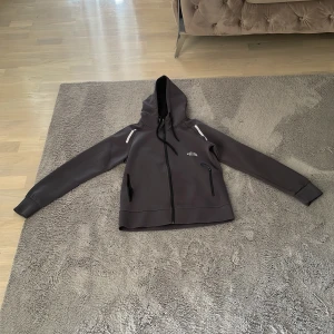 Grå vindjacka från The North Face - Snygg grå vindjacka med huva från The North Face. Jackan har dragkedja framtill, två fickor med dragkedja och svarta detaljer. Perfekt för blåsiga dagar och har en clean, sportig look med diskret logga på bröstet.