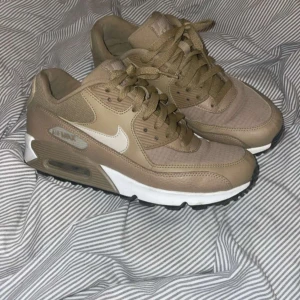 Beige Nike Air Max 90 sneakers - Snygga Nike Air Max 90 sneakers i beige med vita detaljer och klassisk synlig Air Max-sula. Skorna har en mix av skinn och textil, rund tå och snörning. Perfekta för dig som gillar stilrena och neutrala färger med en sportig vibe.